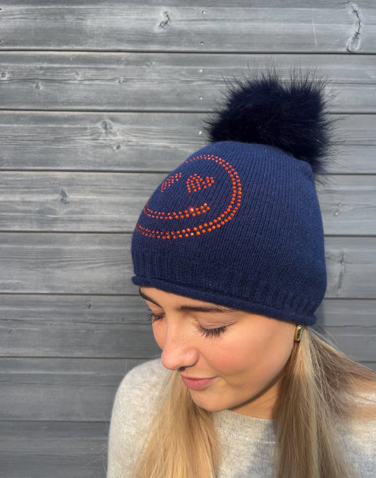 Navy cashmere plain knit beanie with an orange smiley embellishment & faux fur pom